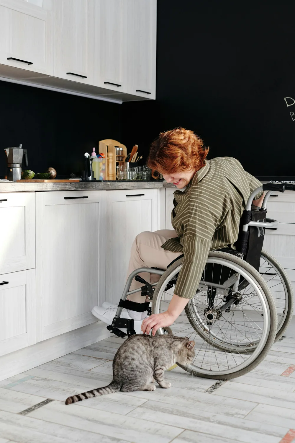 A woman in a wheelchair smiling and patting her cat in a kitchen at home